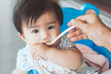 Grandmother feeding cute asian baby girl with a spoon at home. Little baby girl sitting in the chair and eating her meal with innocence.