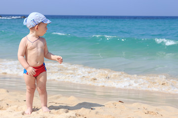 Little baby boy standing on the sand and watching at the sea waves.