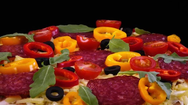 Closeup view of hot homemade pizza with salami and vegetables rotating against black background in 4K. Traditional tasty food.
