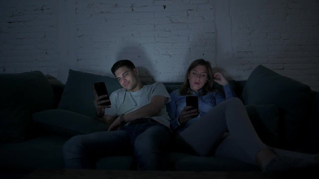 young couple on their 20s using mobile phone ignoring each other sitting at home couch concentrated on internet app in relationship lack of communication and smart phone addiction concept