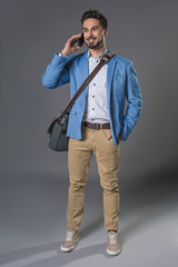 Fototapeta premium Full length portrait of standing attractive happy businessman with crossbody handbag using cellphone