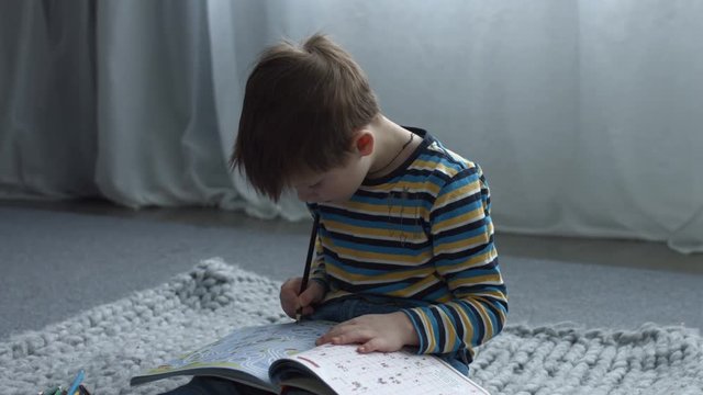Enthusiastic cute preschool kid painting with colorful pencil in coloring book while sitting on the floor in living room. Handsome little boy drawing in child maze book at home.