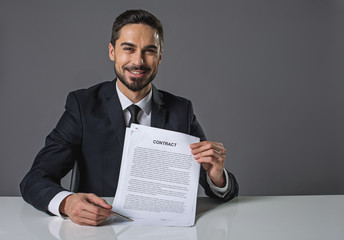 Waist up portrait of young cheerful man is pleased with himself. He is showing a contract and pointing to signature. He looking at camera with happiness. Copy space in right side