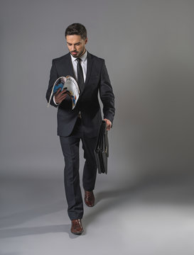 Full length of busy undistracted man in business suit. He is going with briefcase and reading a business periodical