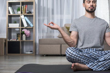 Serene handsome guy relaxing in lotus position in living room. Copy space in left side
