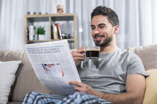 Portrait of contented handsome male reading news on couch and holding cup of coffee