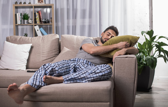 Tranquil young male sleeping on comfortable couch in living room. Bookshelf on background