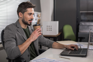 Serene intelligent guy relaxing in front of his office computer and holding paper cup