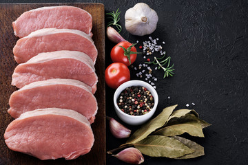 Raw sliced pork loin with tomato, pepper, rosemary, bay leaf and garlic on a dark background. Fresh meat. © vitals