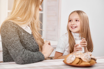 Obraz na płótnie Canvas Smiling little daughter kepping milk glass near mother. There is a big plate full of delicious cookies nearby. She has blonde straight hair and wears white t-shirt. Girl looks happy.