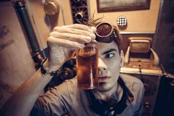 Portrait of a young mad scientist dressed in steampunk style and glasses in his fantastic laboratory with antique bottles.