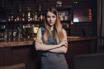 Portrait of young female model with fair hair leaning her elbows on bar counter looking at camera in vintage restaurant