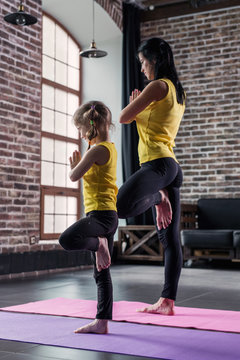 Mother and daughter wearing sports clothing practicing yoga together meditating standing on one leg with hands in prayer position in loft apartment