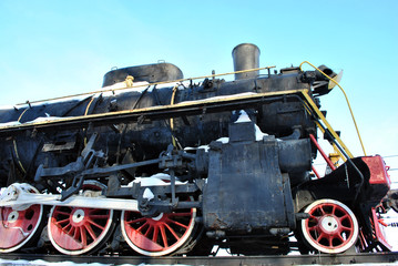 Old black train with red elements covered with snow, standing on platform against blue sky background, close up of side of locomotive, Kharkiv, Ukraine