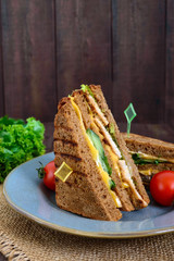Delicious club-sandwich with rye bread, chicken, cheese, cucumbers, greens on a dark wooden background.