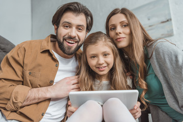 smiling parents and daughter looking at camera and holding tablet