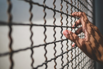 hand of prisoner holding rustic metal fence with pattern shadow, criminal locked in jail waiting...