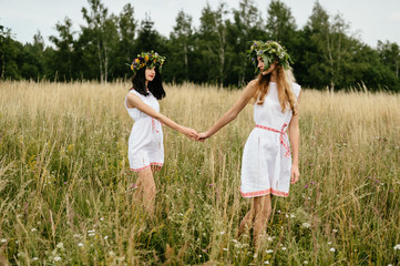 Female friendship. Mood portrait of two beautiful Slavonic appearance young girls in etnic folk dresses and wreath of flowers walking at nature. Tender relationship. Friends leisure. Hand holding hand