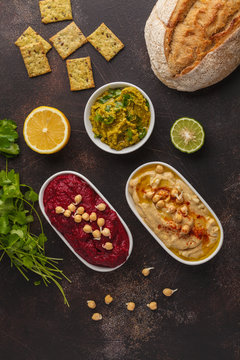 Vegan food background. Vegetarian snacks: hummus, beetroot hummus, green peas dip, vegetables. Top view, dark background, copy space.