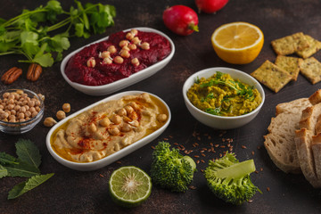 Vegan food background. Vegetarian snacks: hummus, beetroot hummus, green peas dip, vegetables. Top view, dark background, copy space.