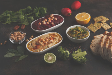 Vegan food background. Vegetarian snacks: hummus, beetroot hummus, green peas dip, vegetables. Top view, dark background, copy space.