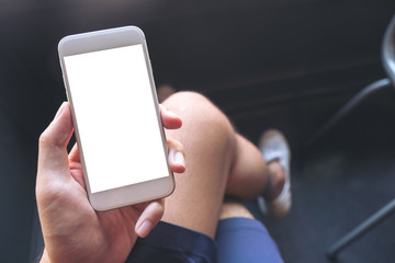 Mockup image of a man's hand holding white mobile phone with blank desktop screen on thigh with white canvas shoes in cafe with feeling relaxed