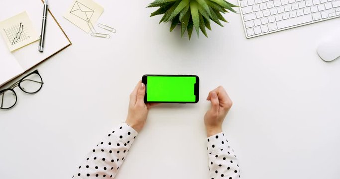 Top view on the white office desk and black smartphone with green screen and female hands taping on it. Horizontal. Office stuff beside. Chroma key.