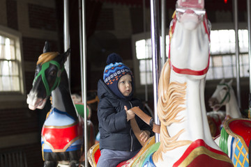 Happy cute toddler in warm clothes riding carousel horse during family trip in New York Central...