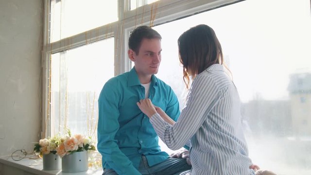 Sunlit young couple sitting by the window and kissing, both in shirts, the girl correcting the guy's tie
