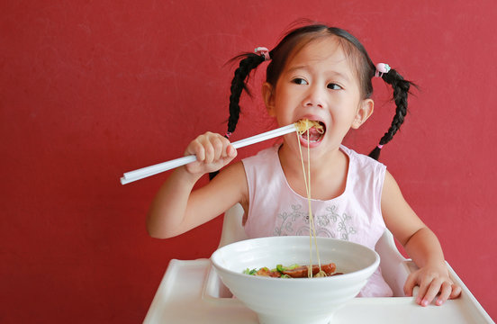 Little asian girl eating Egg noodle with roasted duck and pork on high chair against red wall background.