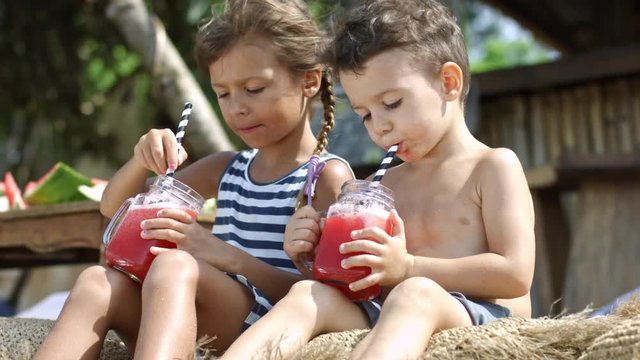 Side view of two children sitting at the beach and drinking watermelon smoothies with drinking straws