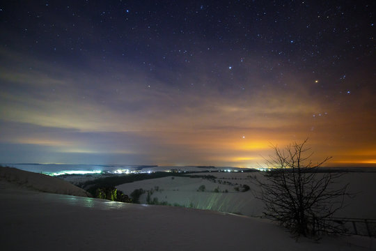 Stars of the night sky are hidden by clouds. Snowy winter landscape at dusk. City in the valley.
