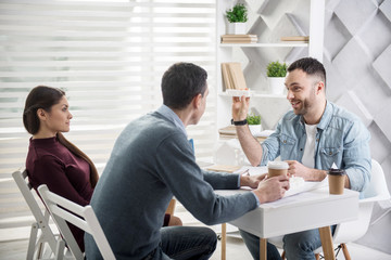 Working time. Handsome cheerful young bearded man smiling and holding a house miniature and having coffee with his colleagues