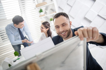 Fototapeta premium Office work. Attractive content young bearded employee smiling and holding a sheet of paper and his colleagues working in the background