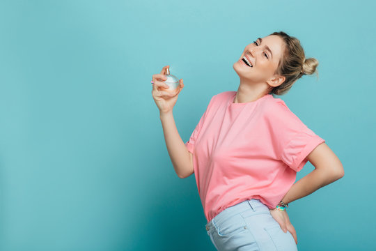 young positive woman holding a perfume bottle and applying it, while standing against blue background. applying fragrance