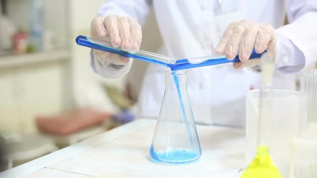 Cinemagraph of unrecognizable scientist wearing rubber gloves adding blue liquid from two test tubes into flask in order to mix them, close-up shot
