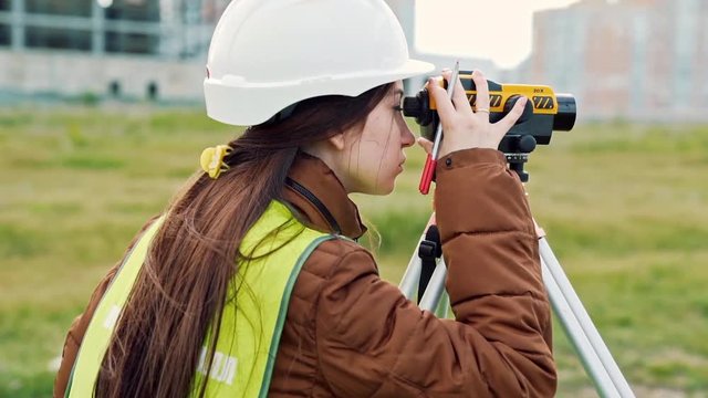 young woman surveyor in green work clothes and helmet adjusts the equipment, produces calculations and writes to the notebook on the construction site. The concept of landscape design, geodesy