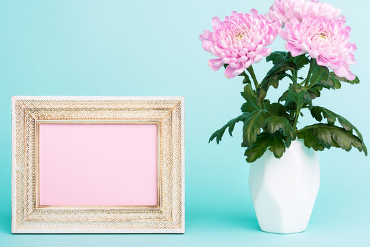 Happy Mother's Day, Women's Day, Valentine's Day or Birthday Pastel Candy Blue Coloured Background. Bright pink flowers in a vase on a table with empty picture frame greeting card.