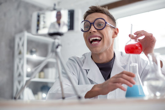 In high spirits. Attractive exuberant fair-haired teenager wearing a uniform and glasses and holding test tubes while doing an experiment