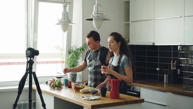 Smiling attractive couple recording video food vlog about cooking haelthy salad on digital camera in the kitchen at home. Vlogging, education and social media concept