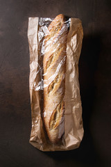 Loaf of fresh baked artisan baguette bread in market paper bag over dark brown texture background....
