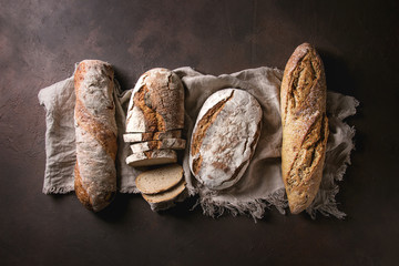Variety of loafs fresh baked artisan rye and whole grain bread on linen cloth over dark brown...