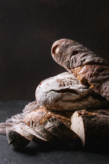 Variety of loafs fresh baked artisan rye and whole grain bread on linen cloth over dark brown texture background. Copy space.