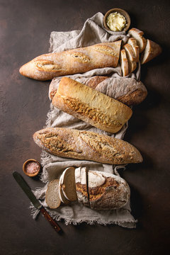 Variety of loafs fresh baked artisan rye, white and whole grain bread on linen cloth with butter, pink salt and vintage knife over dark brown texture background. Top view, copy space.