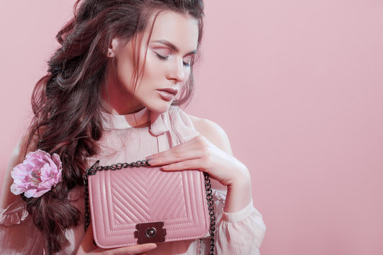 Close up studio portrait of young beautiful woman holding small quilted pink bag. Model with greek braid hairstyle, wearing blouse, posing on pink background. Spring fashion, advertising concept