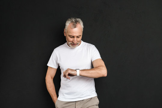 Cheerful mature old man standing isolated looking at his watch.