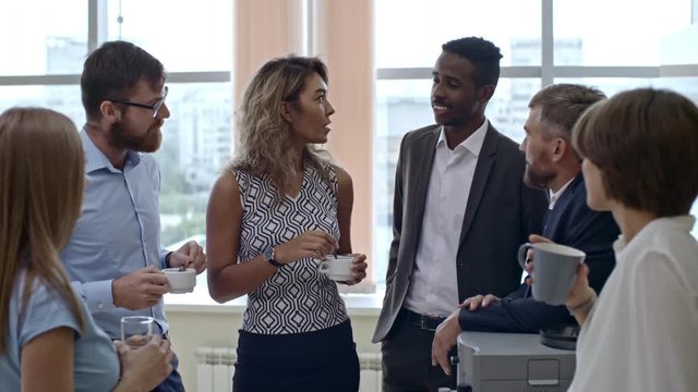 Six colleagues standing together and having conversation over cup of tea when taking break from work