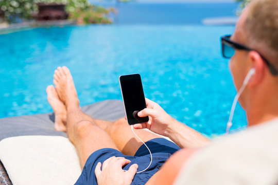 Man sunbathing by the pool and holding mobile phone in hand.