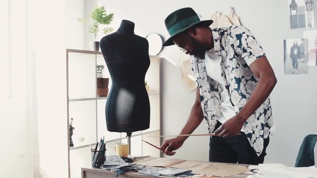 Stylish Afro-American dressmaker in a casual look designing clothes, uses the ruler for his sketches in a modern, cozy office. Professional, creation process. Slow motion, camera stabilizer shot