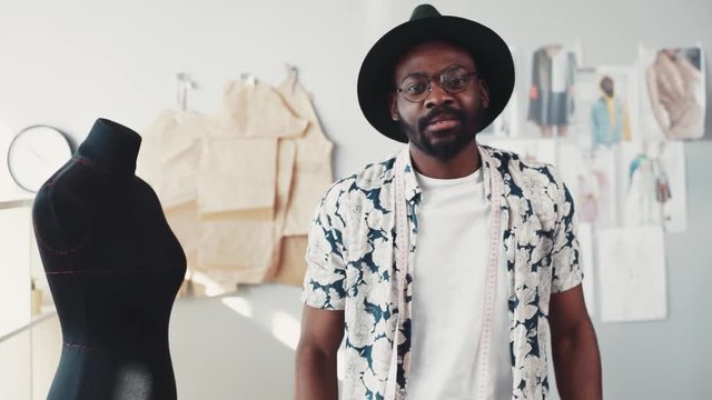 Handsome bearded Afro-American dressmaker in a flowered shirt casually throws the measuring tape around his neck, looks straight to camera. Cozy, modern office. Fashion sketches on the background